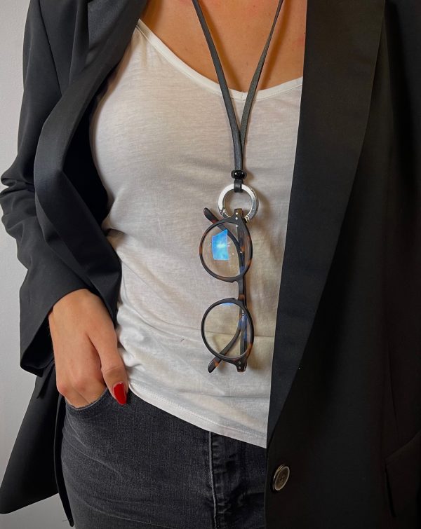 Une dame porte un collier porte lunette en cuir noir avec l'anneau argent, accroché à l'anneau on y retrouve des lunettes